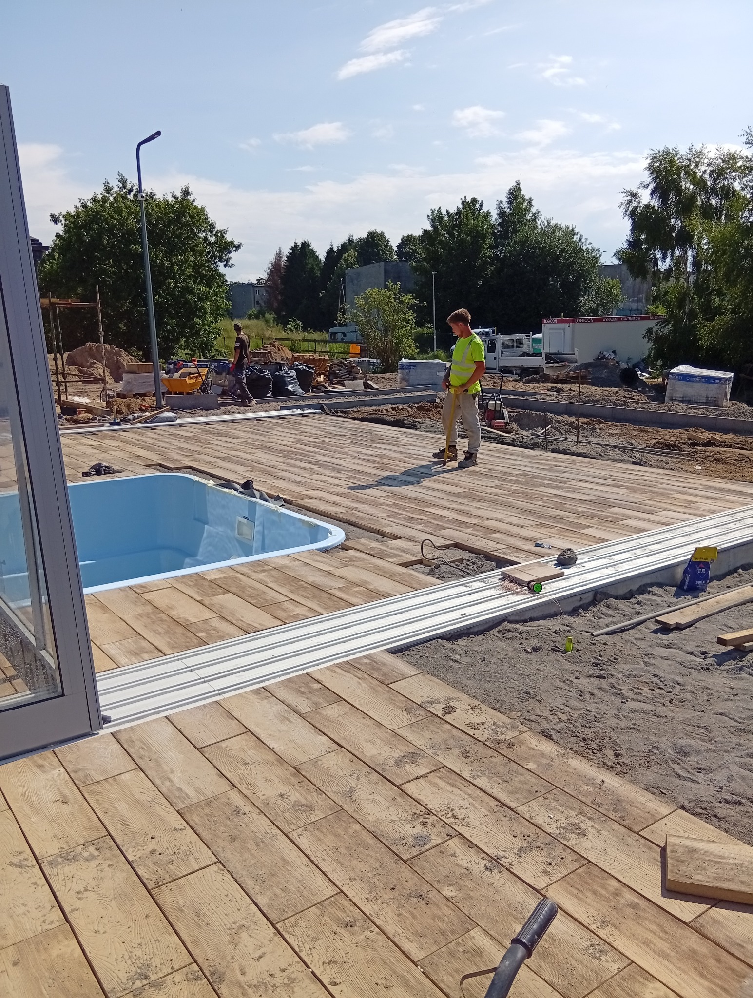 Układanie kostki brukowej imitującej drewno wokół basenu. Pracownik w kamizelce odblaskowej. Widoczne narzędzia i materiały budowlane.
