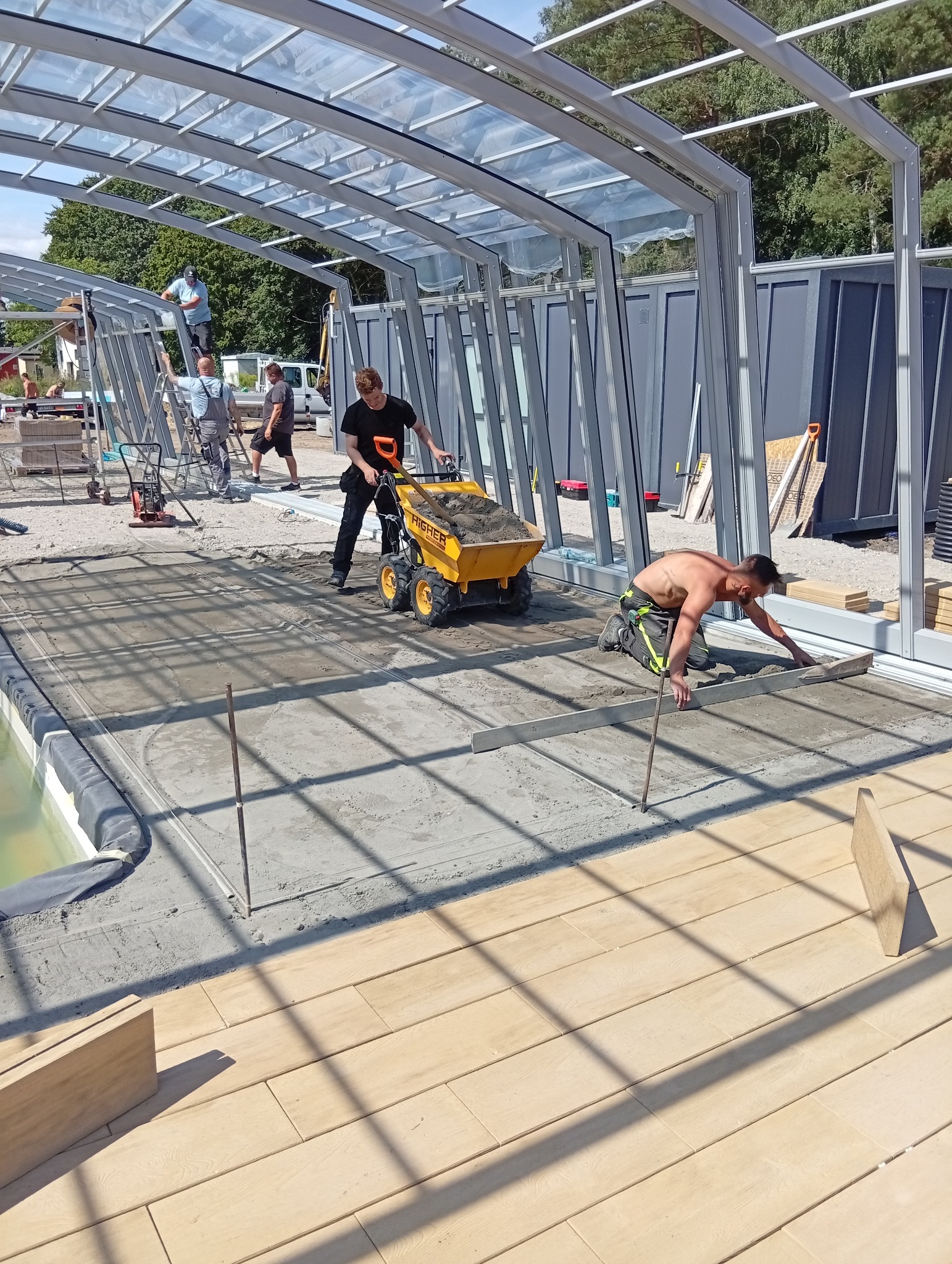 Budowa zadaszenia basenu: pracownicy wyrównują beton, jeden z nich używa mini wywrotki. W tle widoczna konstrukcja szkieletowa i kontenery.