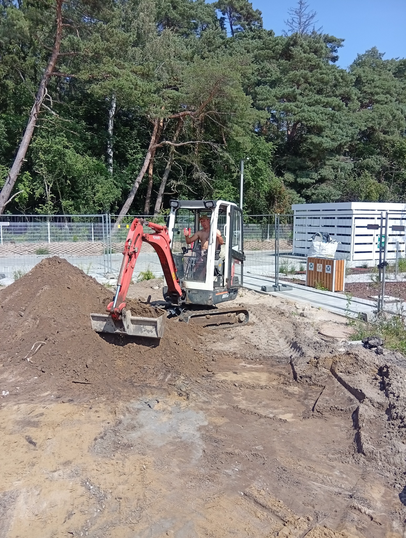 Mini koparka Kubota w trakcie prac ziemnych na terenie zielonym z drzewami w tle, operator w kabinie, widoczne ślady opon i przesypana ziemia.