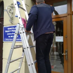 Pracownik na drabinie czyści okno wejściowe do kancelarii radcy prawnego, widoczny spryskiwacz i ściereczka.