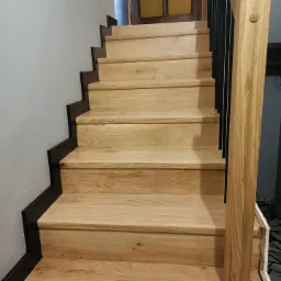 Jasne, drewniane schody z czarnymi elementami wykończeniowymi i balustradą z pionowymi, czarnymi prętami.