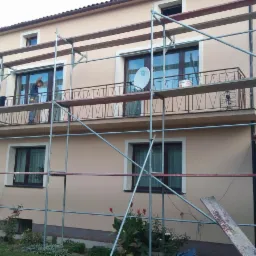 Dom w trakcie prac elewacyjnych. Widoczne rusztowania, pracownik na balkonie i wiadro z farbą. Świeżo pomalowana elewacja w odcieniach beżu.