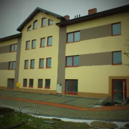 Dwukondygnacyjny budynek z żółtą elewacją i brązowymi pasami, czerwone ramy okienne i drzwi, kostka brukowa przed budynkiem, pochmurne niebo.