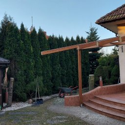 Patryk Pawlik - Konstrukcja drewniana zadaszenia tarasu w trakcie budowy, widoczna taczka i rząd wysokich tuj w tle.