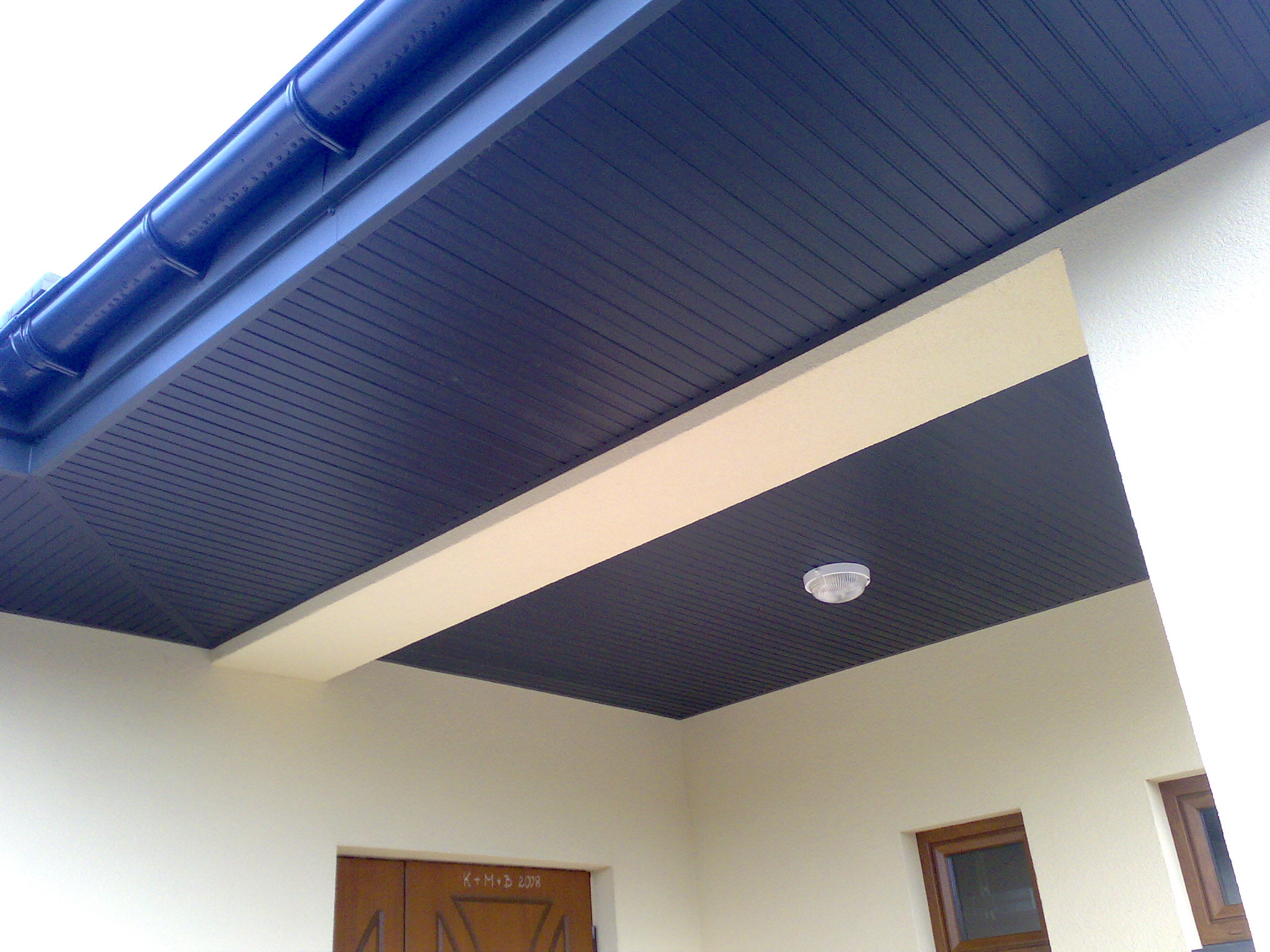 Zamontowana, ciemna podbitka dachowa z oświetleniem, widoczna z dołu, z fragmentem rynny i elewacji budynku.