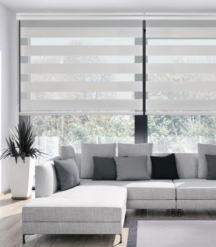 Jasnoszara sofa modułowa z poduszkami w odcieniach szarości i czerni na tle okien z roletami dzień noc w paski, w nowoczesnym salonie z rośliną w białej donicy.