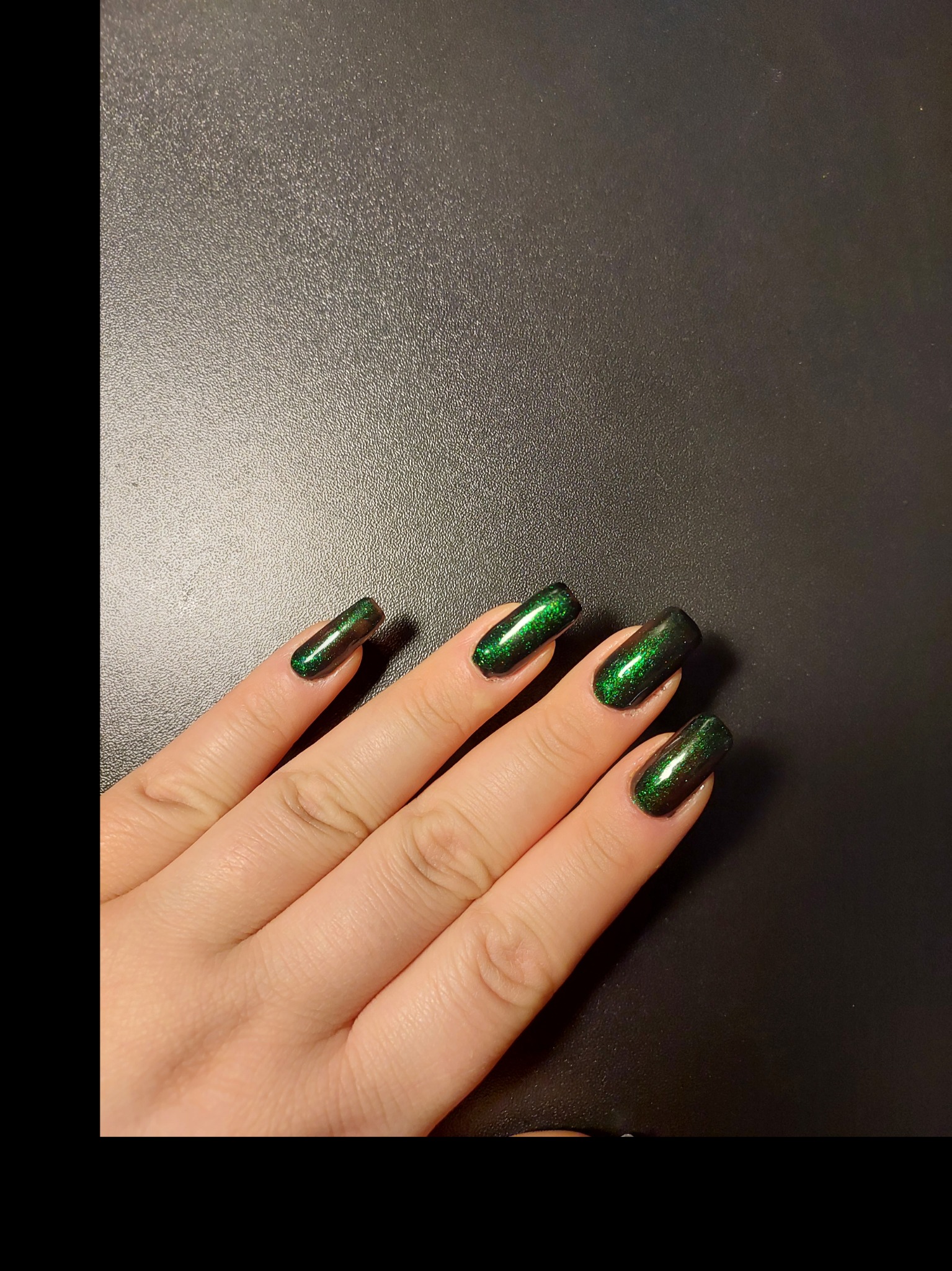 Dłoń z ciemnozielonym, brokatowym manicure na czarnym tle, oświetlenie podkreśla połysk lakieru.