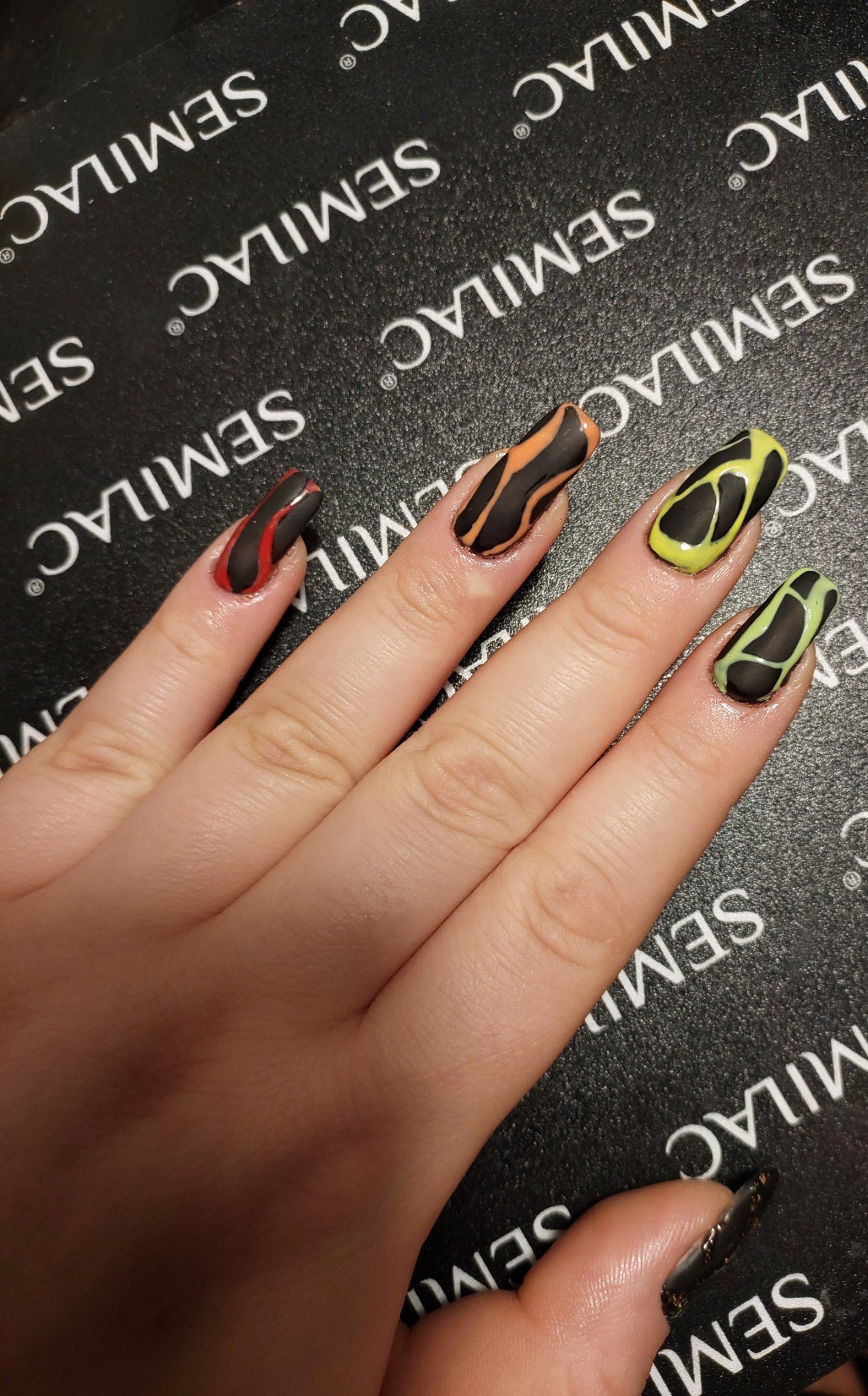 Dłoń z pomalowanymi na czarno paznokciami o wydłużonym kształcie, każdy z innym wzorem w odcieniach czerwieni, pomarańczy i zieleni, na tle podkładki z logo Semilac.