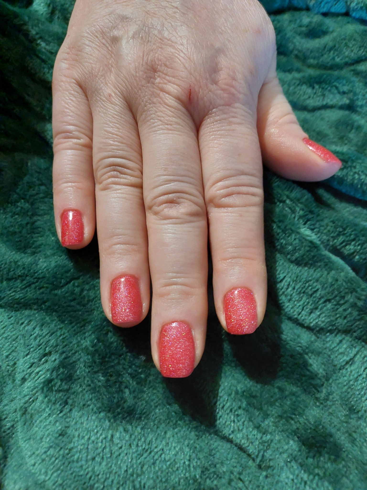 Dłoń z pomalowanymi na czerwono brokatowymi paznokciami na tle zielonego, pluszowego materiału.