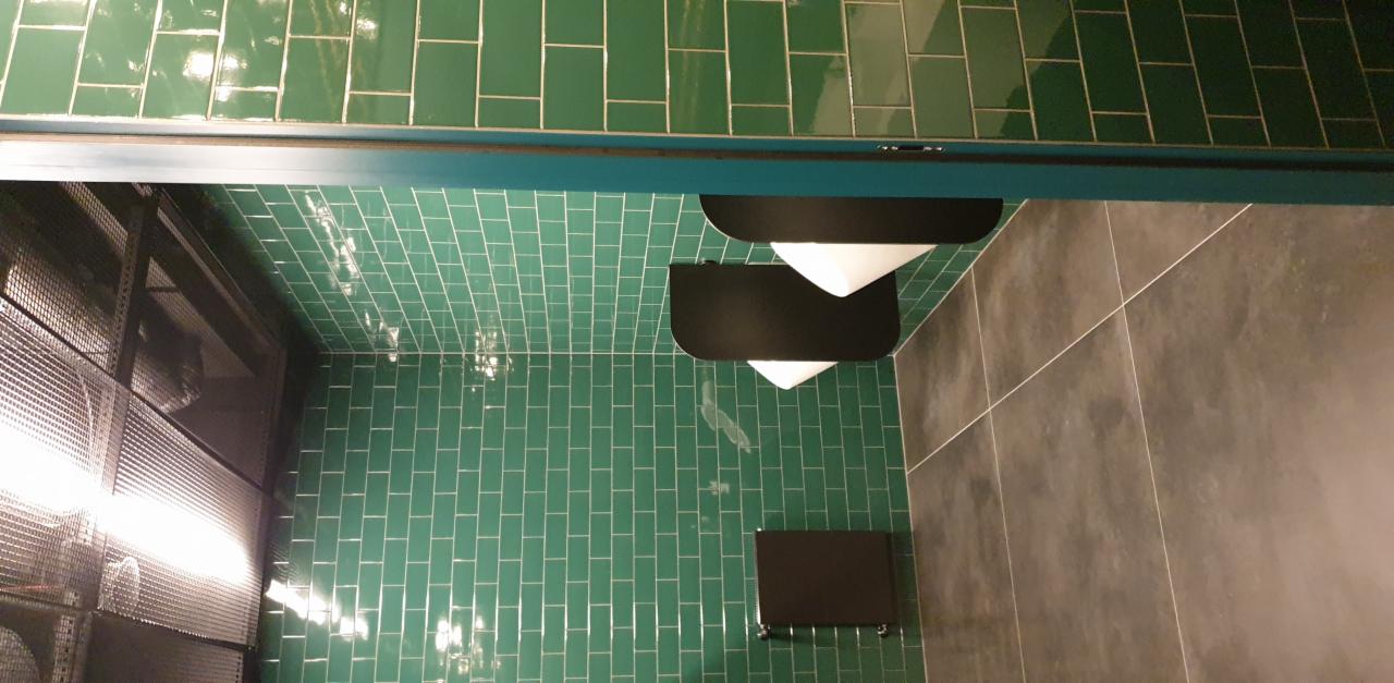 Wykończona łazienka biurowa z zielonymi płytkami typu metro na ścianach i szarymi płytkami podłogowymi, widoczne czarne akcesoria łazienkowe.