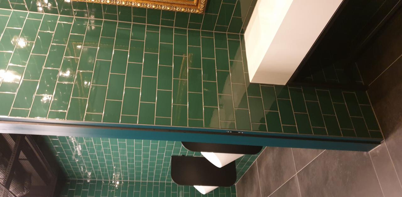 Ściana wyłożona błyszczącymi, szmaragdowymi płytkami w układzie cegiełki, z widocznym fragmentem złotej ramy obrazu u góry i elementami wyposażenia łazienki w dolnej części kadru.