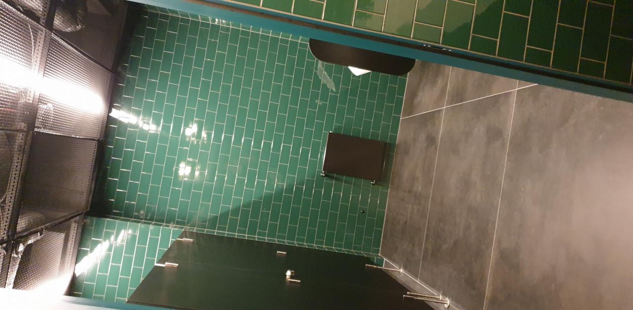 Wnętrze toalety biurowej z zielonymi płytkami typu metro na ścianach, grafitową podłogą i czarnymi kabinami WC.