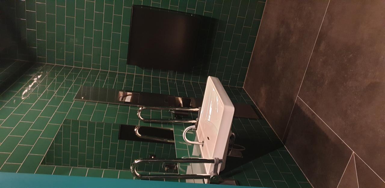 Nowoczesna łazienka biurowa z zielonymi płytkami na ścianach, czarnym panelem nad umywalkami i brązową podłogą z geometrycznym wzorem.