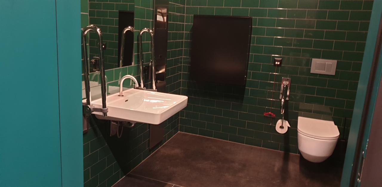 Wykończona łazienka biurowa dla osób niepełnosprawnych z umywalką na regulowanej wysokości, poręczami i toaletą na tle ciemnozielonych płytek.