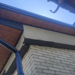 Widok z dołu na fragment budynku z nową, ciemną rynną i drewnianą podbitką dachową, z widocznym ociepleniem ściany i fragmentem starej rynny w tle.