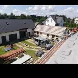 Prace dekarskie w Baćkowicach: Widok z góry na dach w trakcie krycia, z widocznym samochodem dostawczym, stertą łat i blach, oraz dwoma dekarzami pracującymi na dachu. W tle budynki mieszkalne...