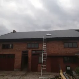 Dwukondygnacyjny budynek z czerwonej cegły z nowym szarym dachem z blachy trapezowej i oknem dachowym, na dachu stoi drabina oparta o ścianę.