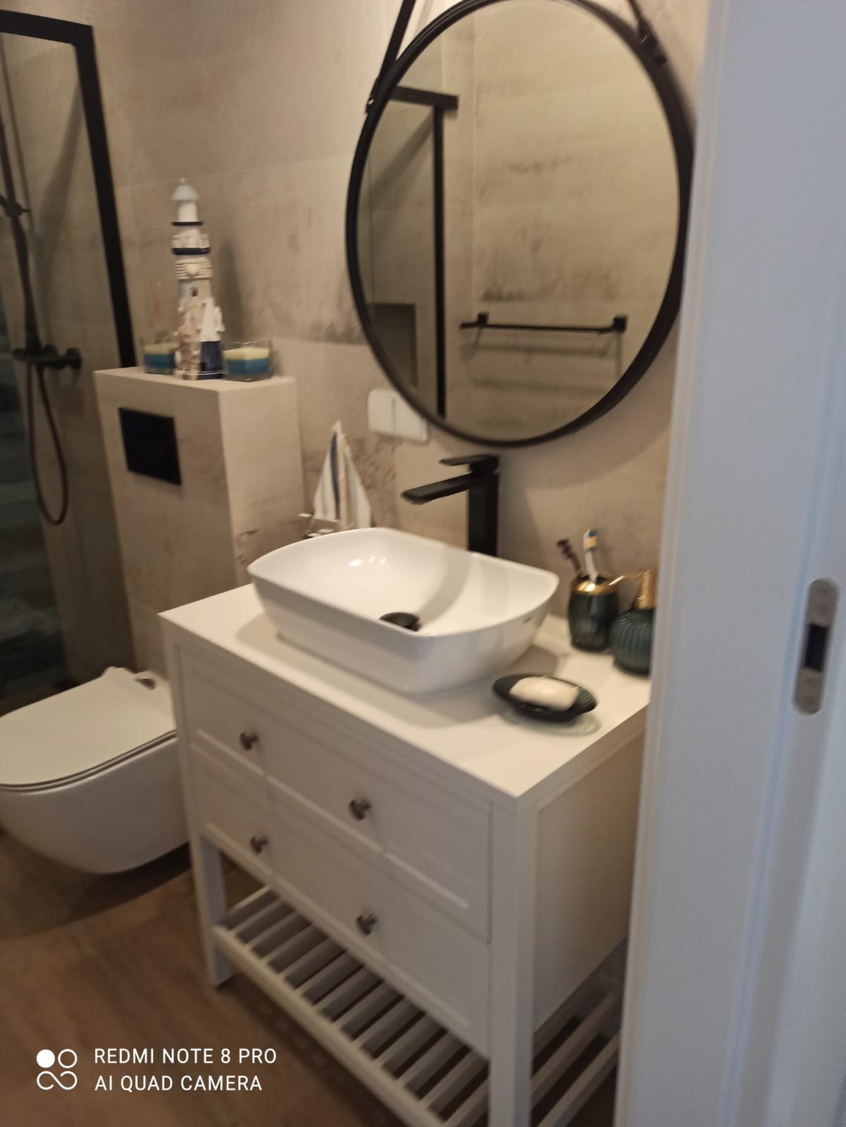 Biała umywalka nablatowa z armaturą w kolorze czarnym, okrągłe lustro w czarnej ramie i dekoracje marynistyczne w łazience w Borówcu, z widoczną toaletą i elementami wykończenia w szarościach.