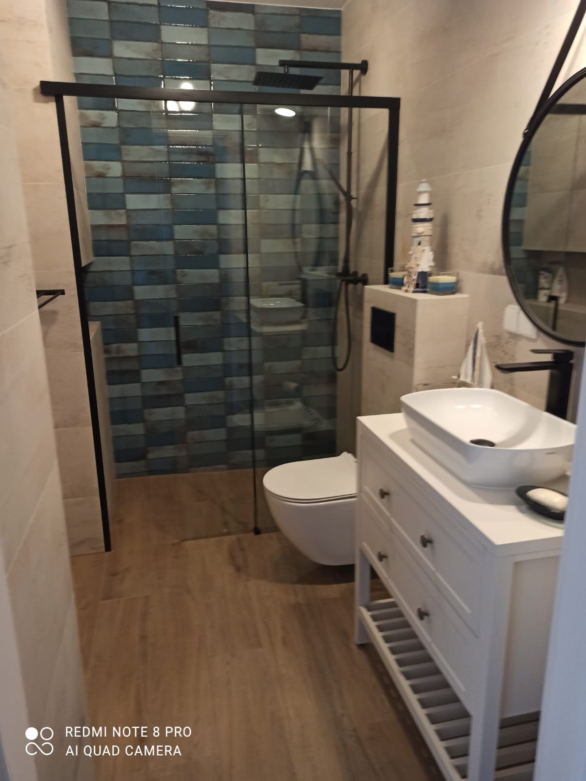 Nowoczesna łazienka z kabiną prysznicową, wykończona płytkami w odcieniach błękitu, z czarną armaturą i białą szafką pod umywalkę w stylu marynistycznym, uzupełniona o dekoracyjny model latarni...