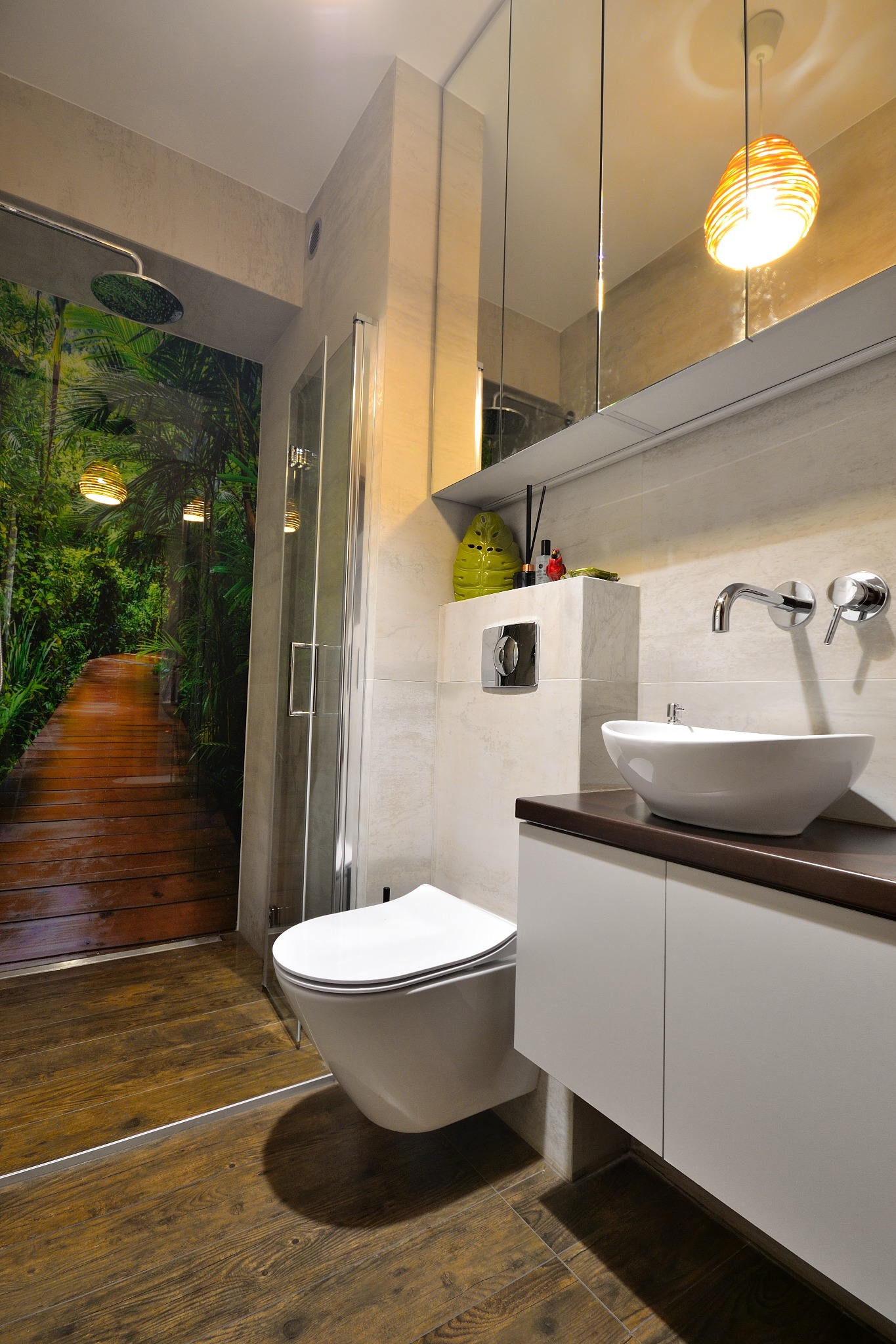 Nowoczesna łazienka z kabiną prysznicową z fototapetą leśną, podwieszaną toaletą i umywalką nablatową na szafce z ciemnym blatem, oświetlona dekoracyjną lampą.