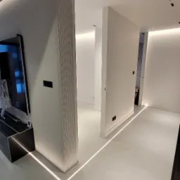 Nowoczesne wnętrze z minimalistycznym oświetleniem LED wzdłuż podłogi, białe ściany i drzwi, telewizor zamontowany na ścianie z dekoracyjnymi pionowymi panelami.