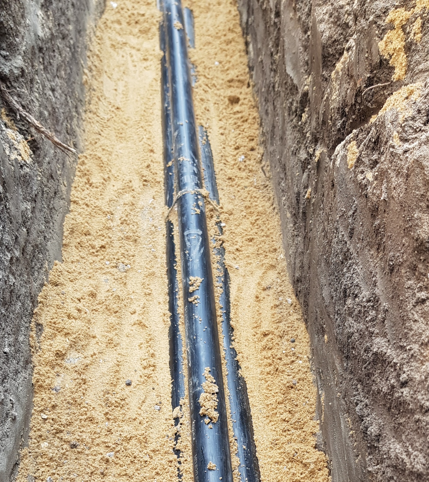 Wykop z trzema czarnymi rurami zabezpieczonymi żółtym piaskiem, widoczne ślady po wykopie ziemi po bokach.