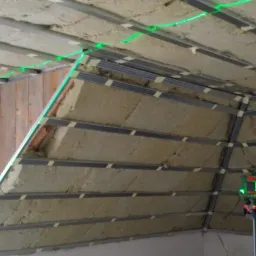 Ocieplenie poddasza wełną mineralną, widoczne profile metalowe, poziomica laserowa wyświetlająca zielone linie, etap prac wykończeniowych.