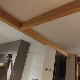 Wnętrze z drewnianymi belkami stropowymi, widoczna kuchnia z szarymi szafkami, ceglana ściana z kominkiem i wiszące lampy w stylu industrialnym.