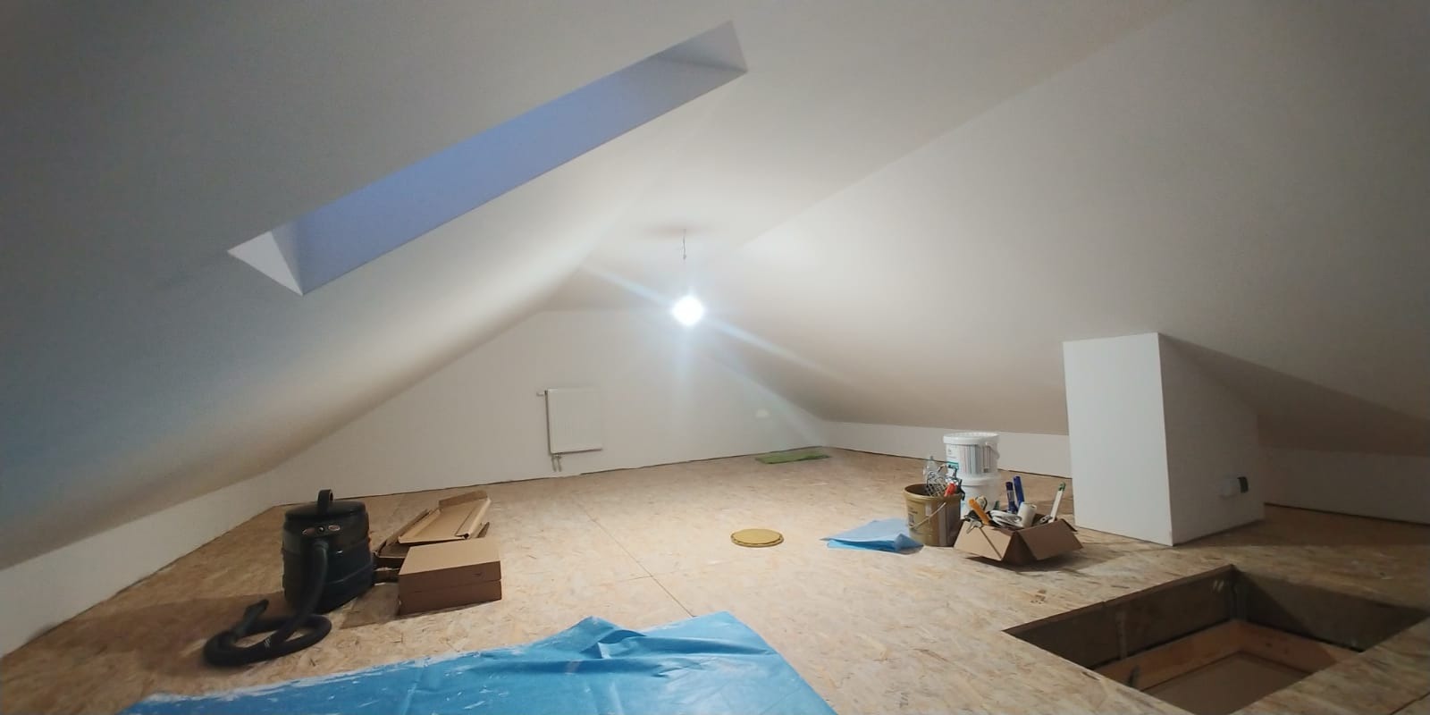 Poddasze w trakcie adaptacji, z widocznym oknem dachowym, częściowo wykończonymi ścianami, podłogą z płyt OSB, narzędziami i materiałami budowlanymi, oraz zabezpieczeniem folią malarską.