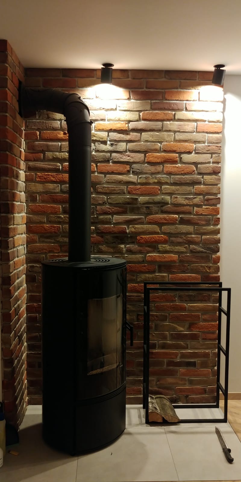 Czarny piec wolnostojący z rurą dymową na tle ściany z cegły dekoracyjnej, oświetlony trzema punktowymi lampami sufitowymi, obok metalowy stojak na drewno i nóż na podłodze z płytek.