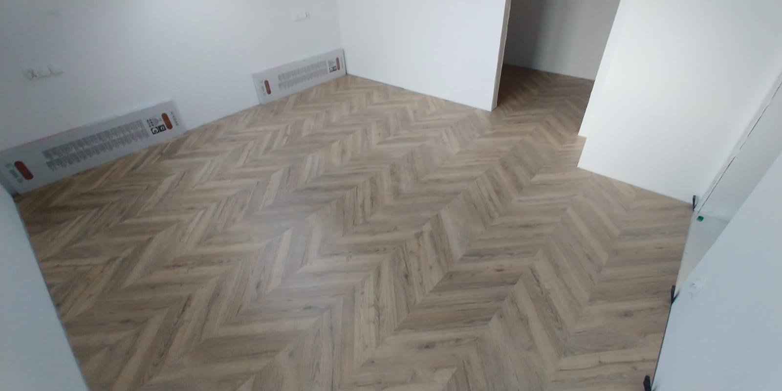 Ułożone panele podłogowe w jodełkę w pustym pomieszczeniu z białymi ścianami i grzejnikiem przypodłogowym.