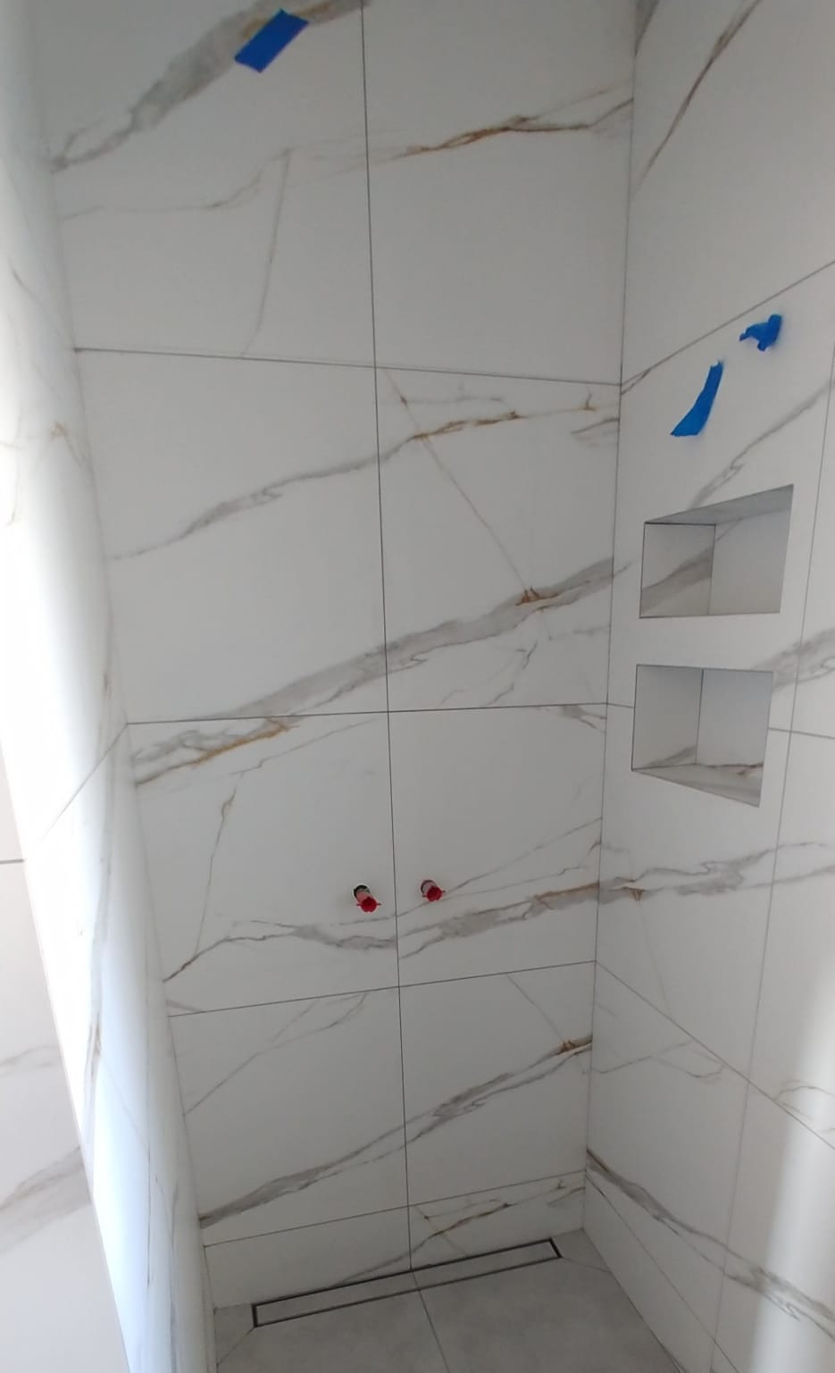 Wykończona kabina prysznicowa z białymi płytkami imitującymi marmur, z wnękami na kosmetyki i odpływem liniowym w podłodze, zabezpieczona niebieską taśmą malarską.