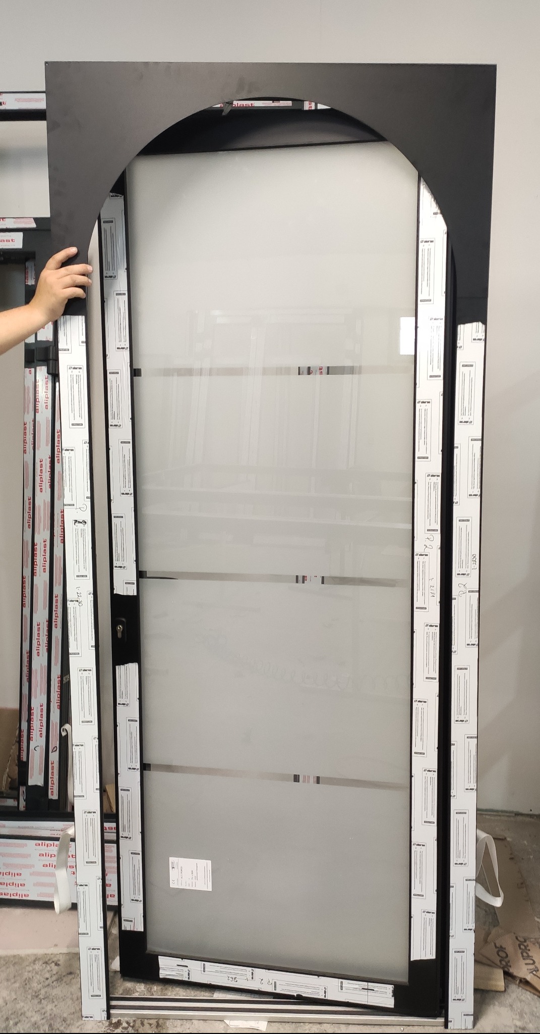 Częściowo zmontowane, czarne, wewnętrzne drzwi z łukowatą górną ramą, z matowym szkłem, oświetlone sztucznym światłem, widoczne profile aluminiowe i naklejki producenta.