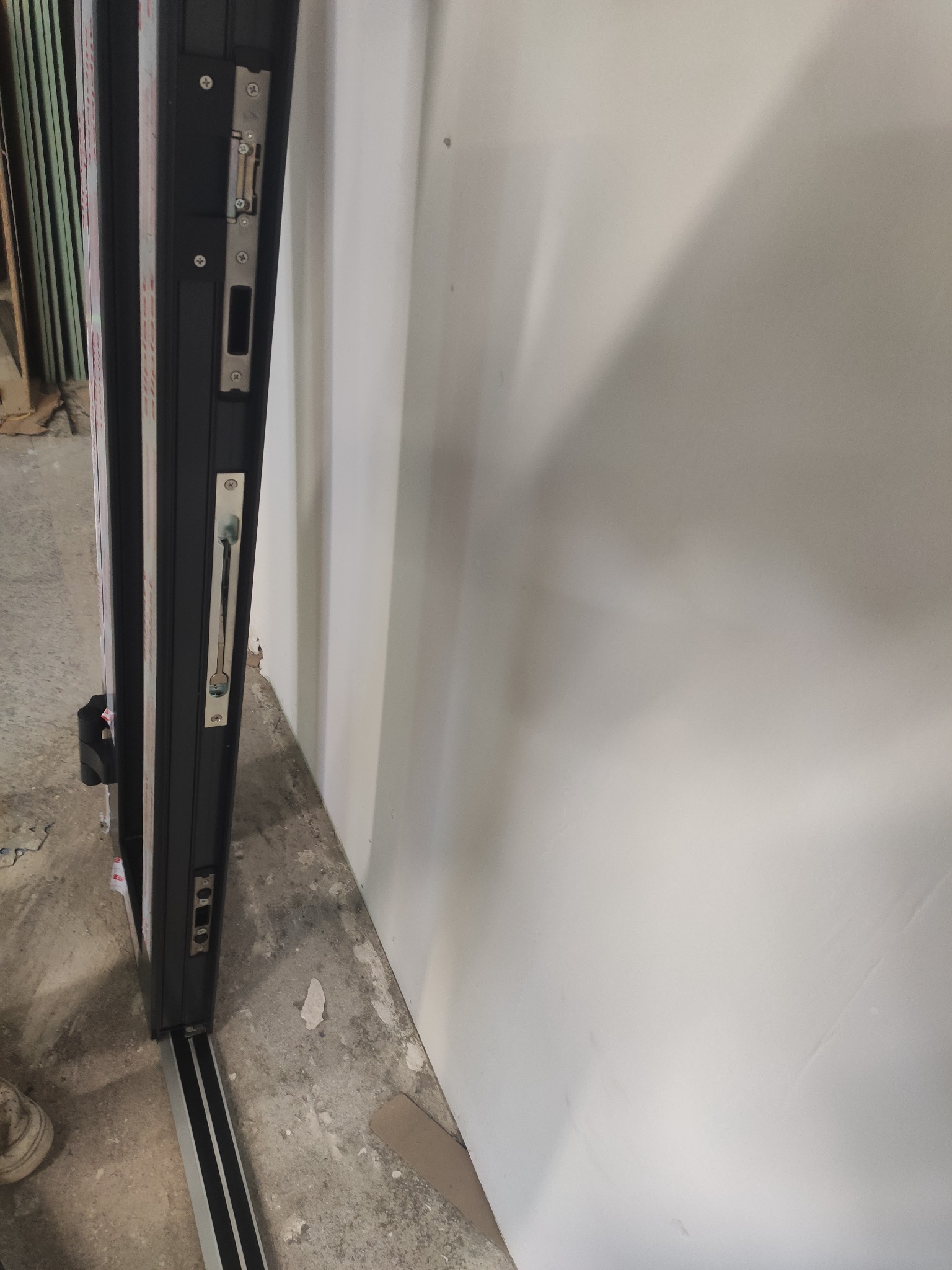 Detal czarnej ramy drzwi przesuwnych z widocznym mechanizmem zamka i prowadnicą dolną, na tle białej ściany i surowej podłogi.