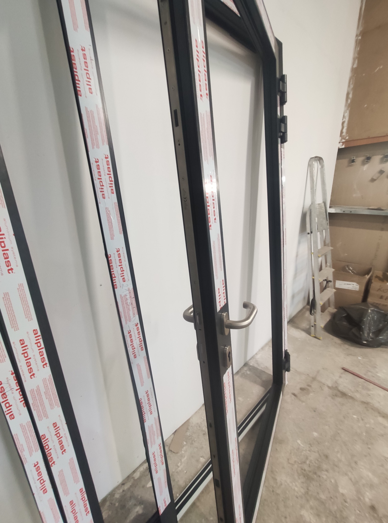 Częściowo zmontowane czarne ramy okienne z naklejkami producenta, widoczne zawiasy i klamka, w tle drabina i kartony w surowym wnętrzu.