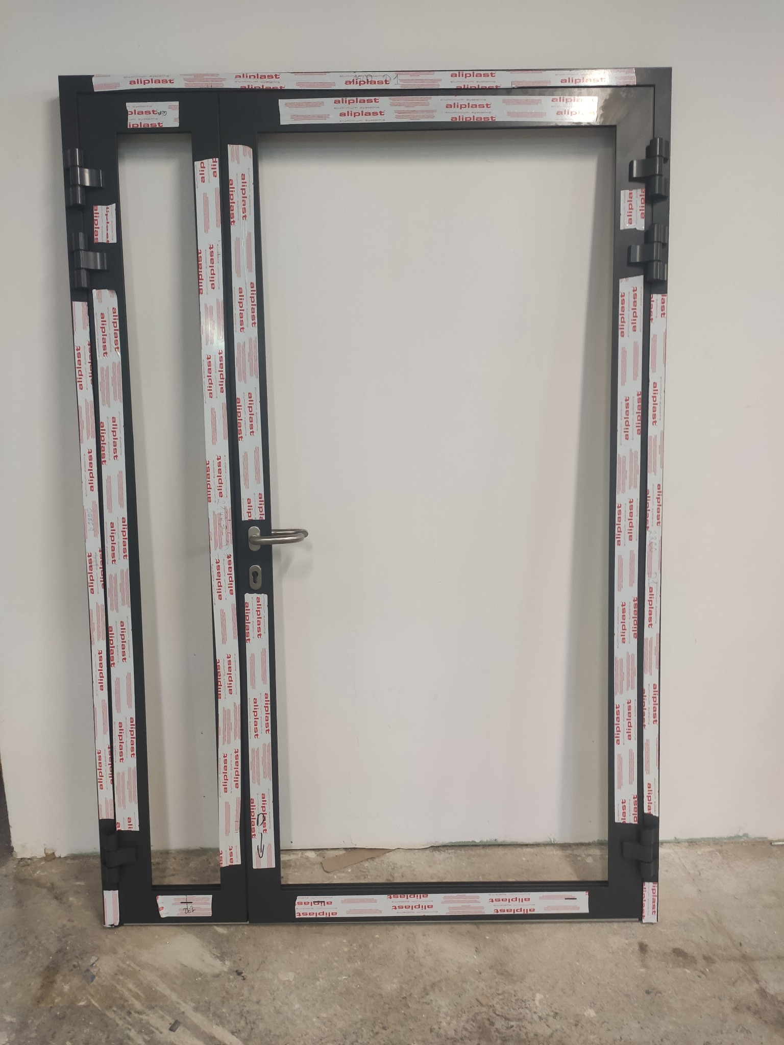 Ciemna rama drzwiowa z zamontowaną klamką, zabezpieczona folią ochronną z nadrukiem producenta, widoczna na tle białej ściany i surowej podłogi.