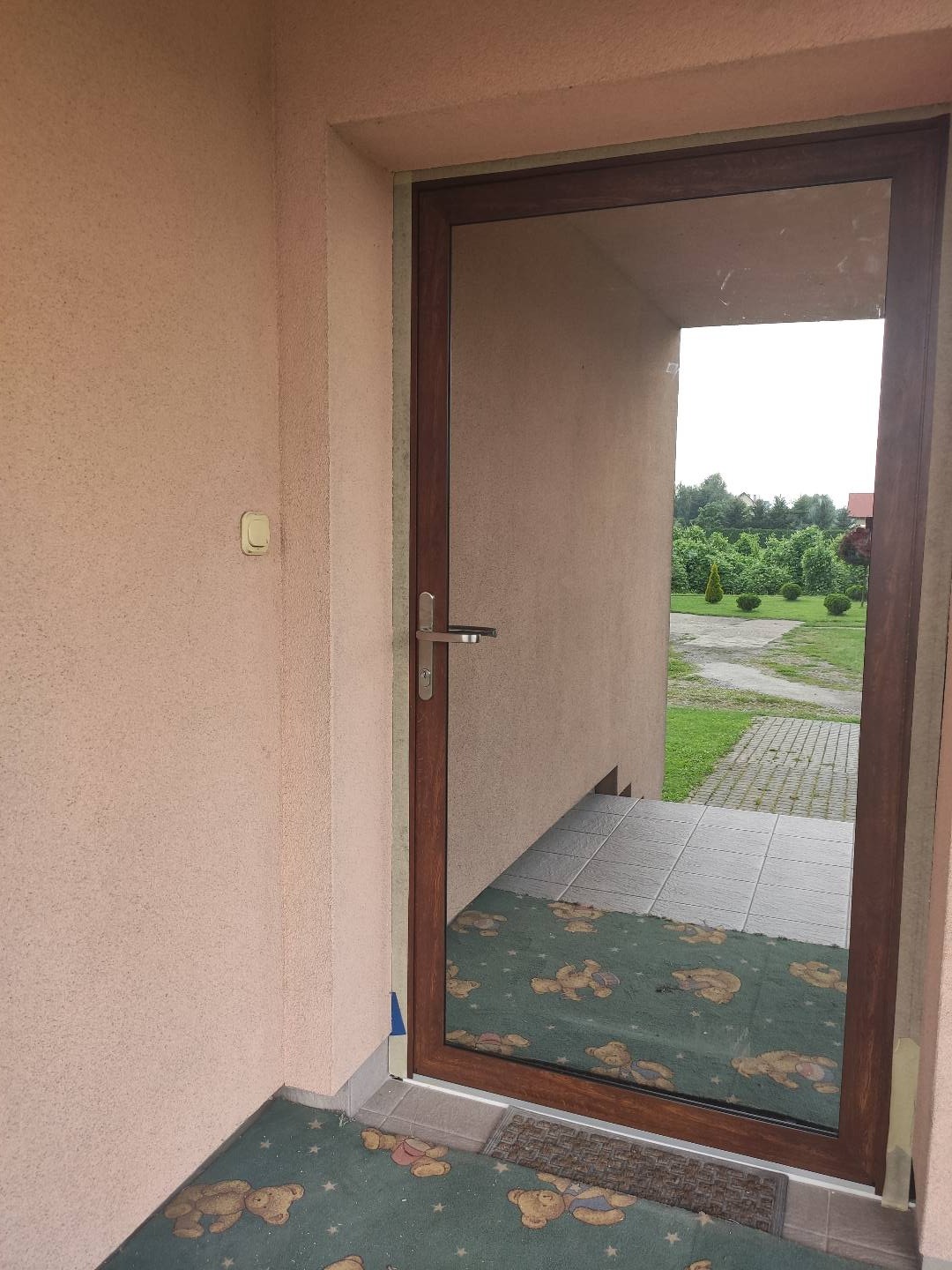 Brązowe drzwi wejściowe z widocznym odbiciem dywanu z motywem pluszowych misiów w szkle, zewnętrzne otoczenie z kostką brukową i zielenią w tle.