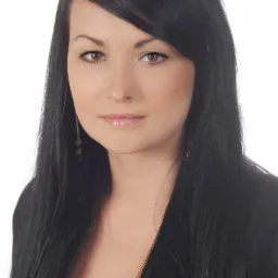 Portret kobiety z brązowymi oczami i długimi, czarnymi włosami, ubrana w ciemną marynarkę na białym tle.