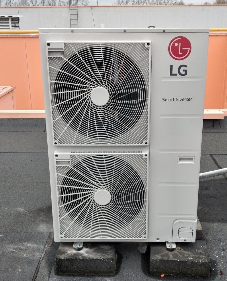 Biała zewnętrzna jednostka klimatyzacji LG Smart Inverter, umieszczona na szarym, płaskim dachu, wsparta na betonowych bloczkach, z pomarańczową ścianą w tle.