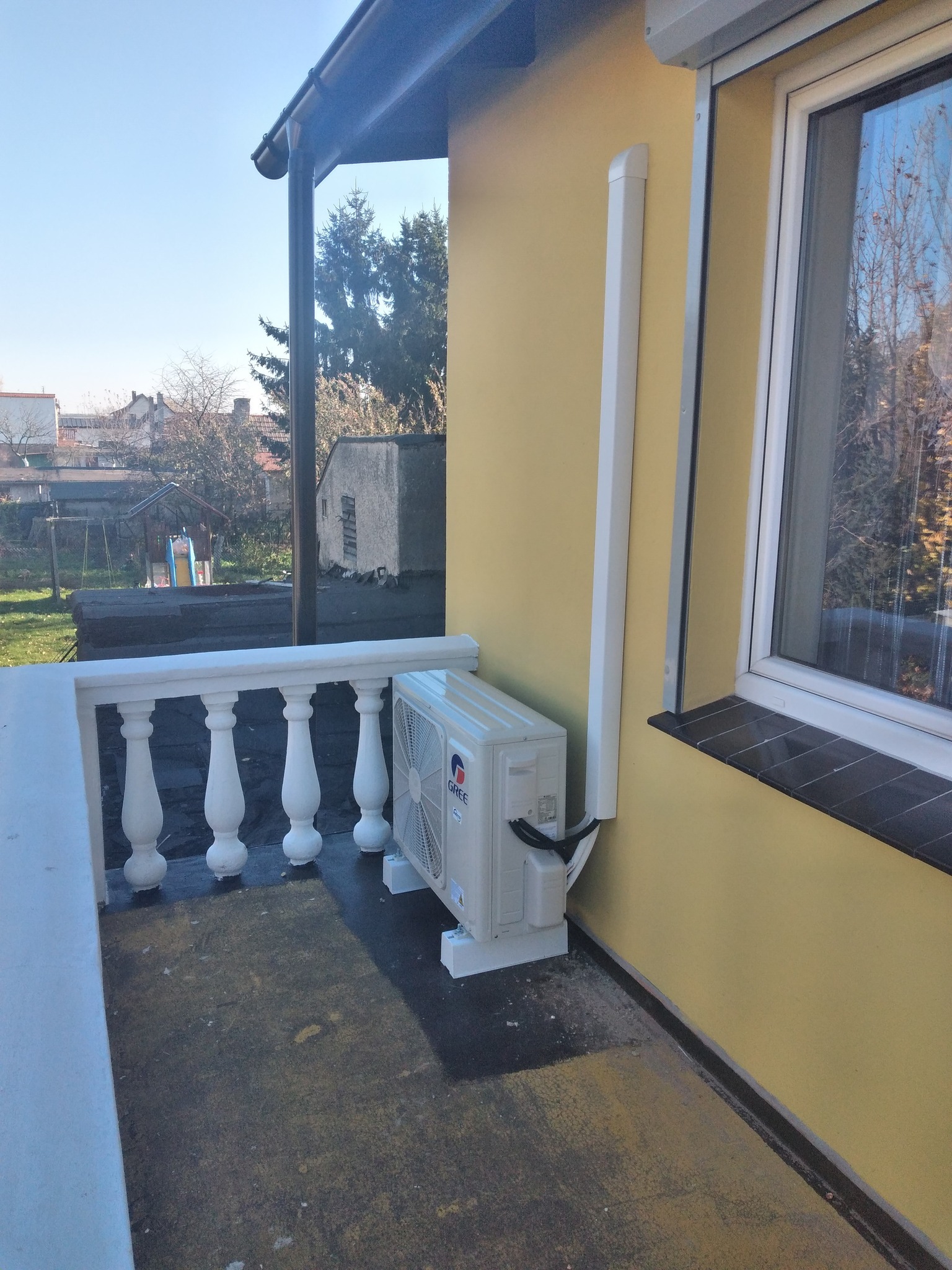 Zewnętrzna jednostka klimatyzacji GREE zamontowana na balkonie budynku o żółtej elewacji, z widoczną białą listwą maskującą instalację.