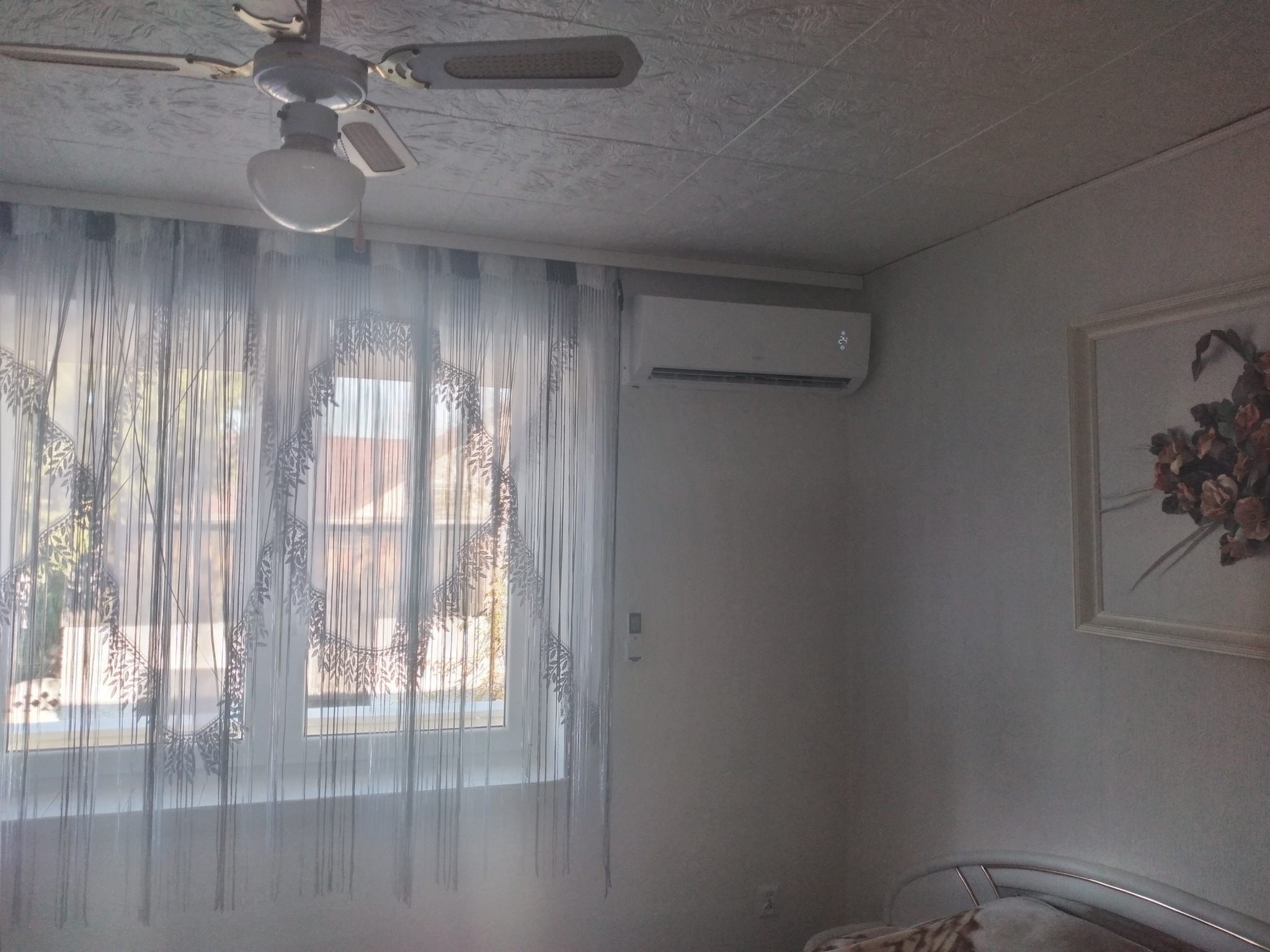 Biały klimatyzator ścienny z wyświetlaczem, zamontowany w pokoju z dekoracyjnymi zasłonami, wentylatorem sufitowym i obrazem w ramie.