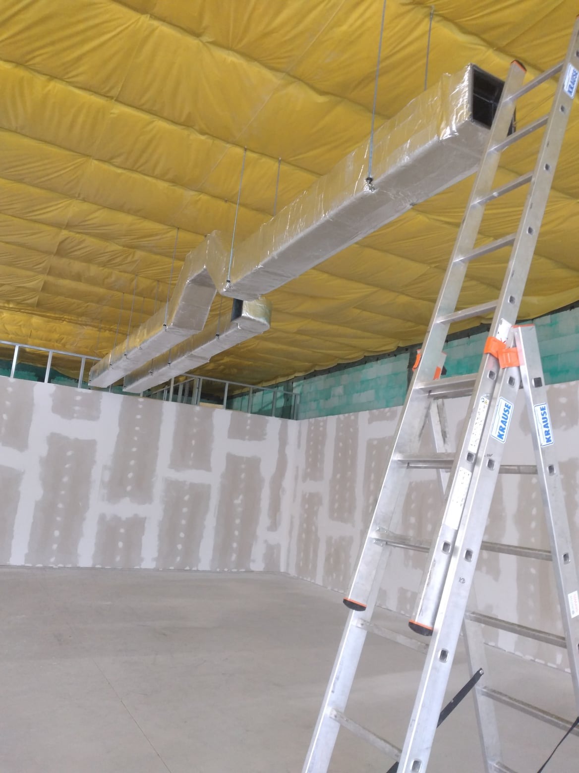 Montaż kanałów wentylacyjnych owiniętych folią na suficie z żółtą izolacją, widoczna aluminiowa drabina Krause i ściany w trakcie szpachlowania.