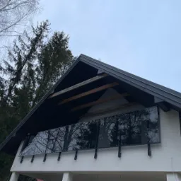 Szklana balustrada na balkonie domu jednorodzinnego w Zegrzu, z widocznymi odbiciami drzew na szkle. Czarna konstrukcja dachu i biała elewacja.