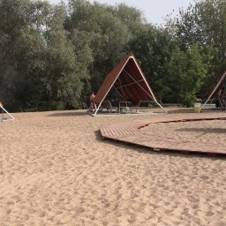 MSC GROUP SP. Z O. O. - Plaża z drewnianymi altanami o trójkątnej konstrukcji, połączonymi ścieżką z desek. W tle zieleń drzew i krzewów. Elementy drewniane, piasek, letni dzień.