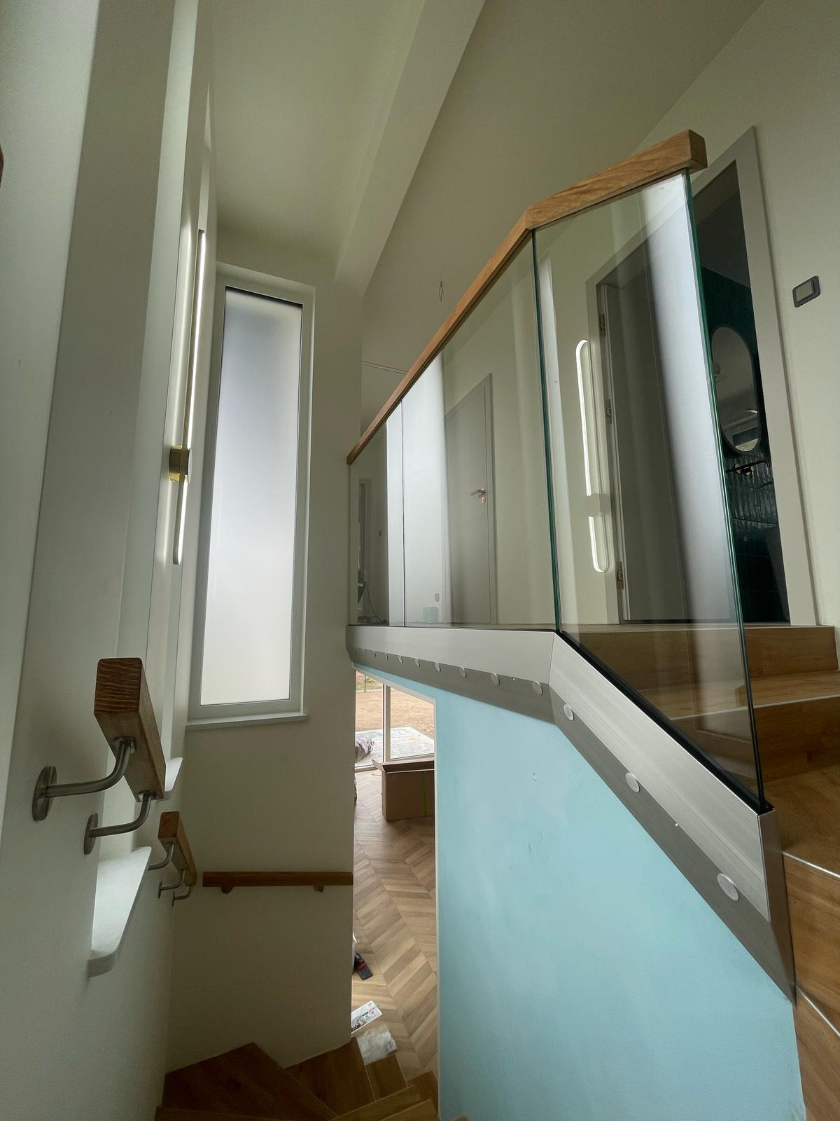 Wnętrze z drewnianymi schodami i szklaną balustradą z drewnianym wykończeniem. Widoczne okno i fragment podłogi z parkietem w jodełkę.