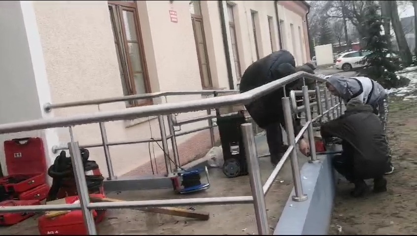 Montaż balustrady przy wejściu do budynku szpitala w Legionowie. Trzech monterów przy pracy, widoczne narzędzia i walizki narzędziowe.
