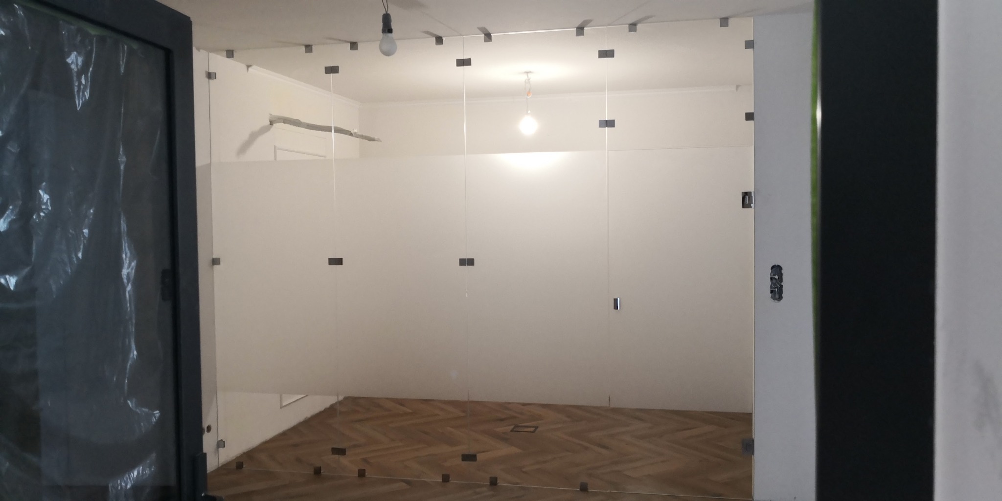 Wnętrze biura w trakcie montażu ścian szklanych z widocznymi mocowaniami. Parkiet na podłodze, surowy stan wykończenia, widoczne kable i niedokończone elementy.