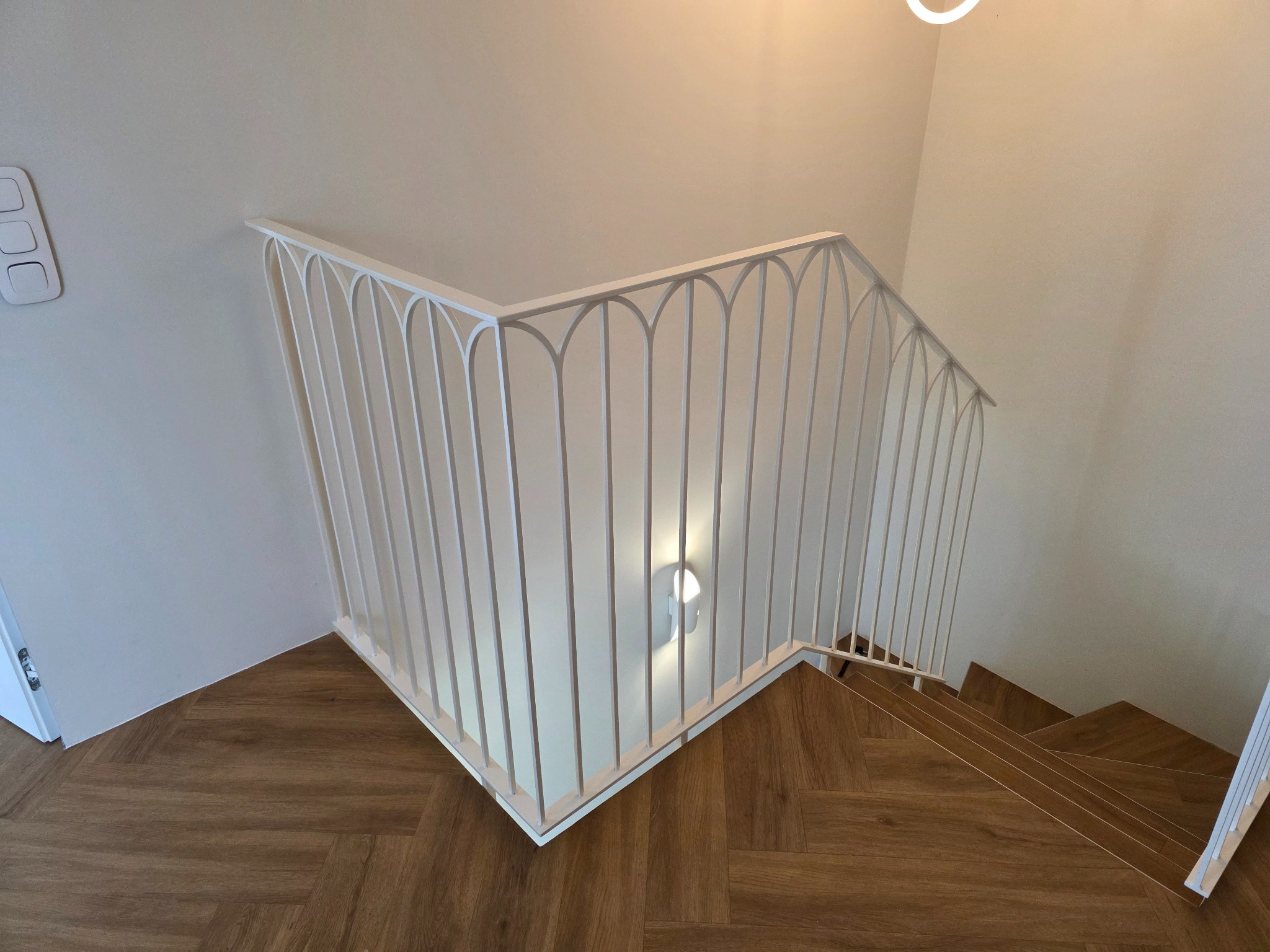 Biała balustrada schodowa z pionowymi prętami w kształcie łuków, drewniane schody i parkiet ułożony w jodełkę, widok z góry.