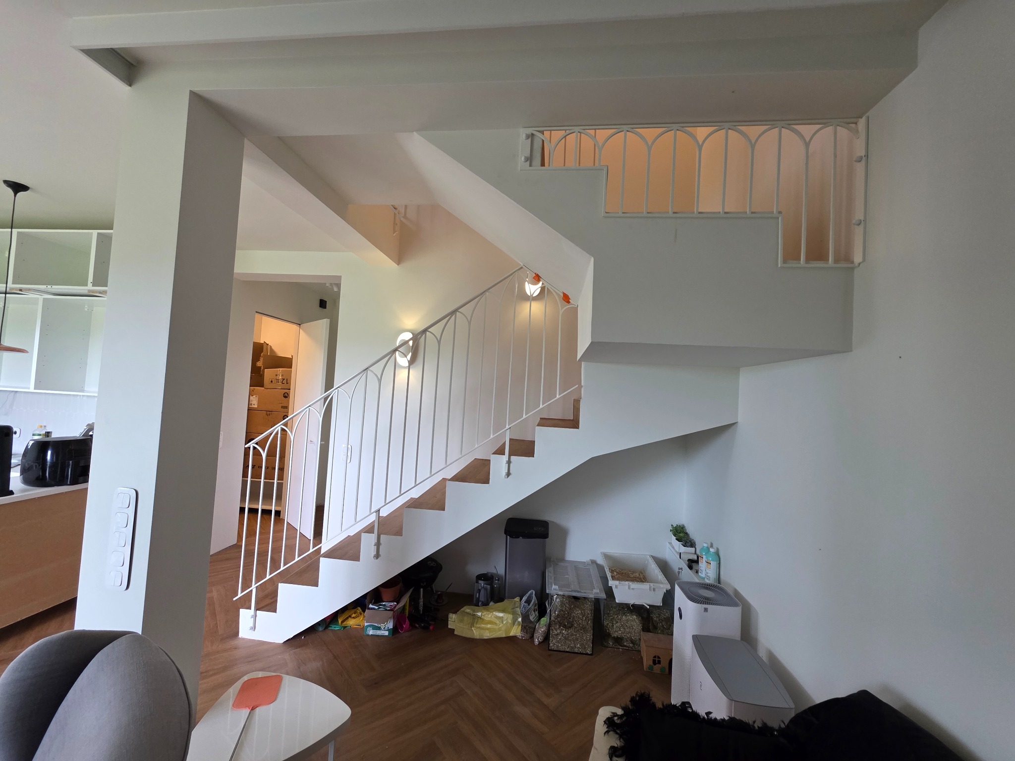 Białe schody z metalową, giętą balustradą w nowoczesnym wnętrzu. Widoczne drewniane stopnie i nietypowe oświetlenie ścienne. W tle elementy wyposażenia mieszkania.