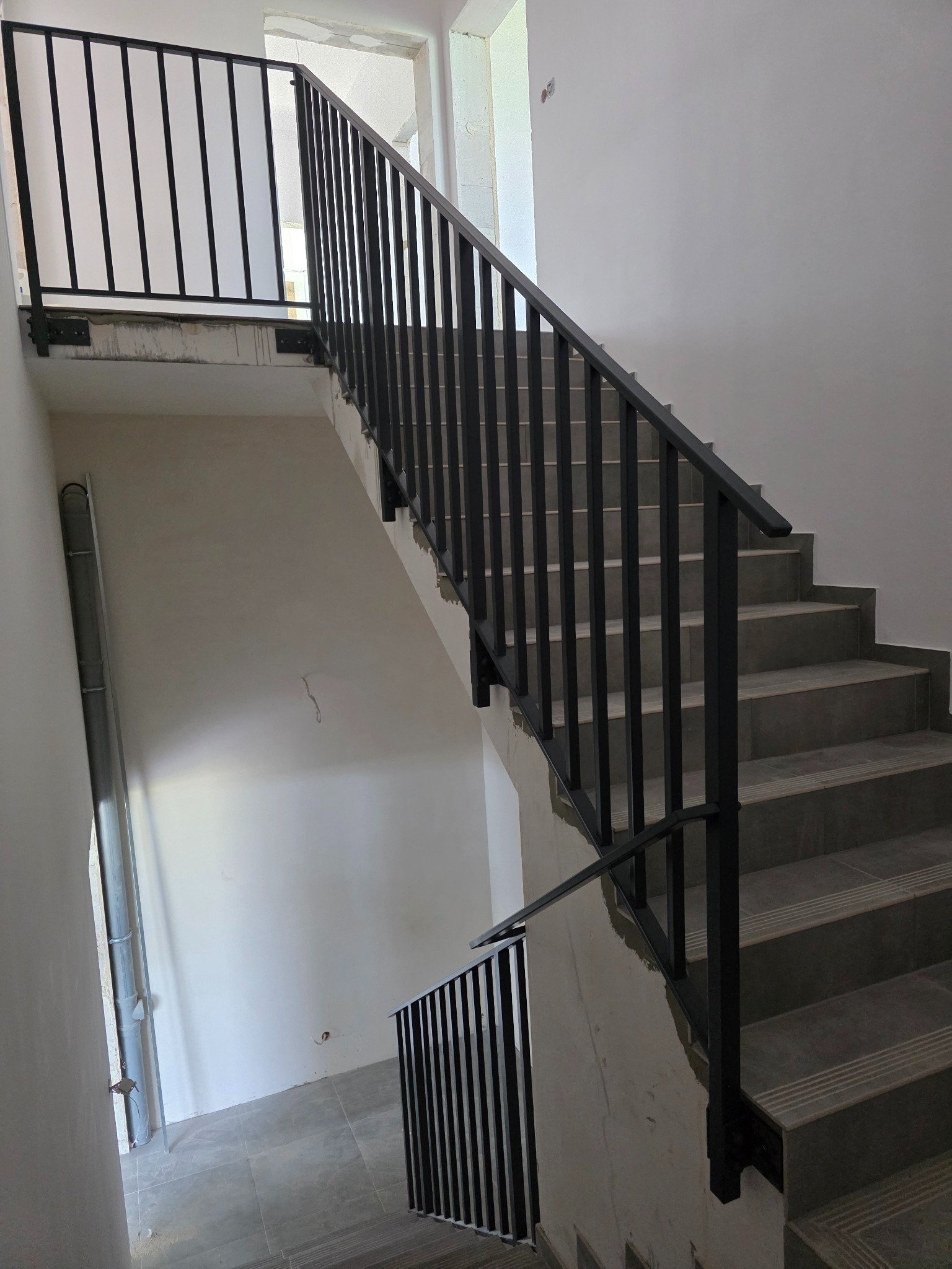 Czarne, metalowe barierki schodowe w surowym wnętrzu. Widoczne szare stopnie i fragment instalacji na ścianie. Ujęcie z dołu, podkreślające pionowe linie.